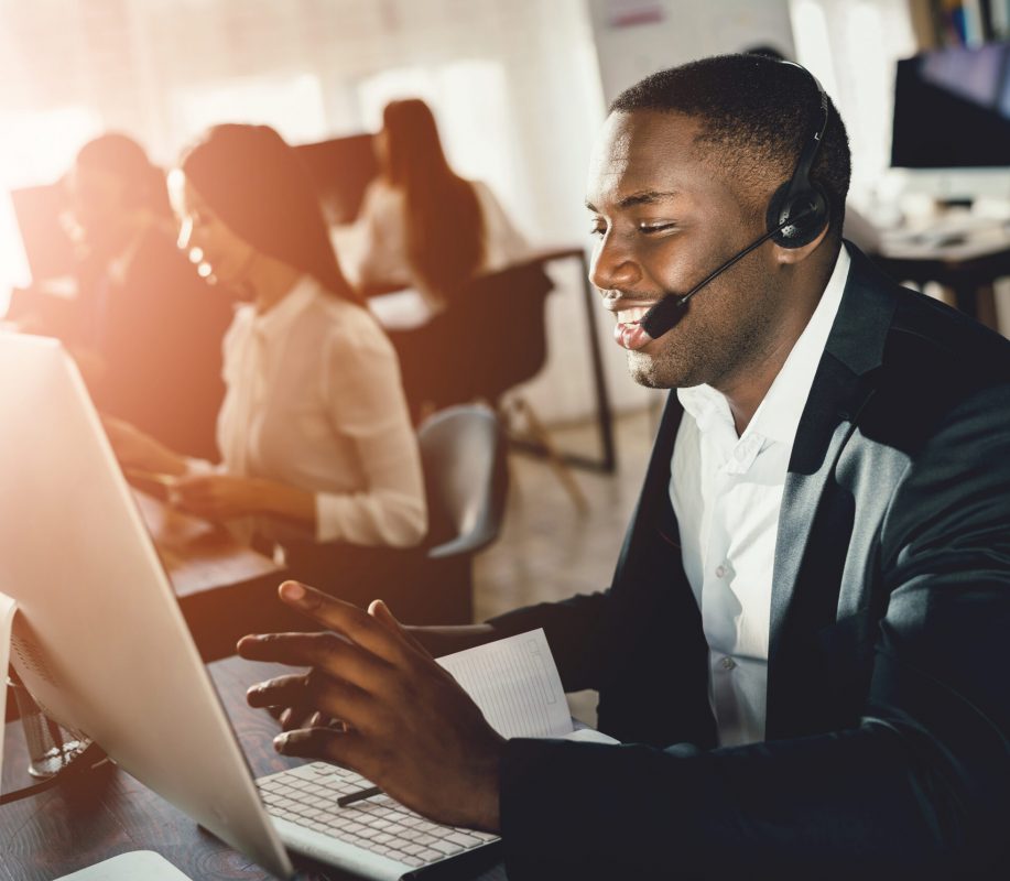 A black guy works in a call center. He has headphones on which they talk with customers. He is an operator and he answers questions.